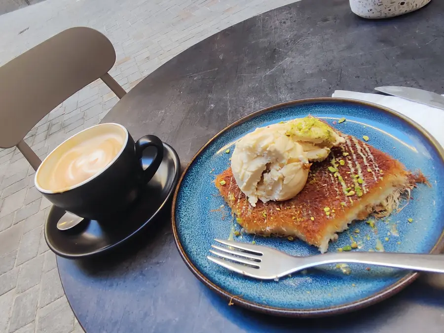 Künefe mit Eis auf blauem Teller und ein Cappuccino auf einem Tisch vor dem Café